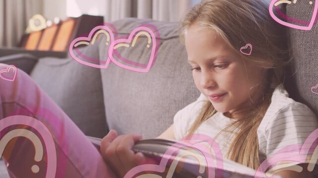 Vertical video: Schoolgirl reading storybook on gray sofa, turning pages as pink hearts appearing