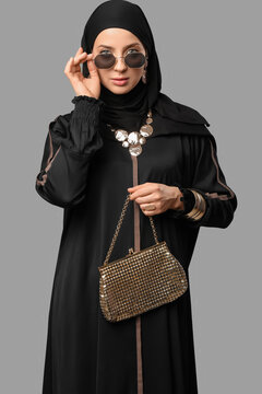 Beautiful young stylish Muslim woman in black hijab with golden accessories and sunglasses on grey background