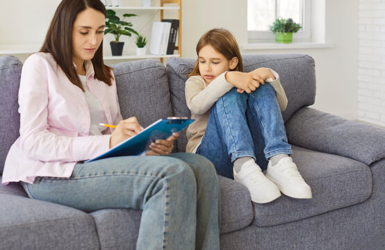 Psychologist meeting with child for mental health counseling and psychotherapy session in home office. Woman writing notes during session on sofa, friendly conversation of child and therapist