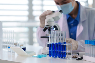 Fototapeta premium Adult Asian female scientist researcher conducts scientific analysis experiment with microscope test tubes blue liquid in modern laboratory setting chemical analysis development
