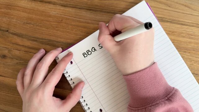 Close up of hands writing &ldquo;BBQ shopping list&rdquo; on lined notebook paper, summer grilling and grocery planning concept, natural light overhead shot on wooden table, 4K video