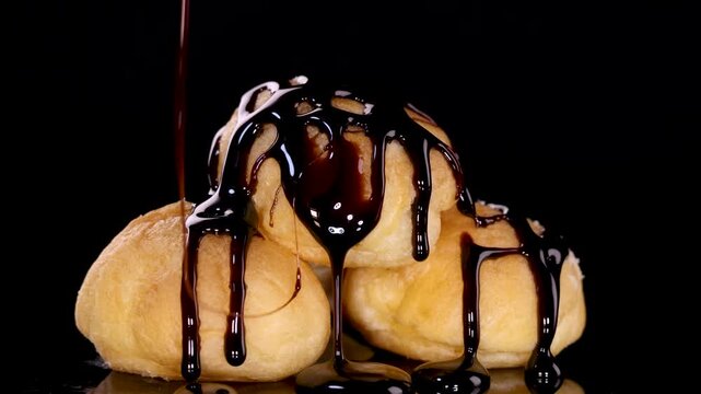 Dark liquid chocolate sauce pouring over delicious cream puffs in slow motion against black background.