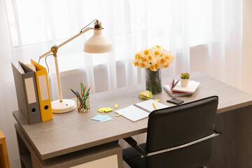Stylish workplace with lamp, bouquet of narcissus flowers and notebook in office