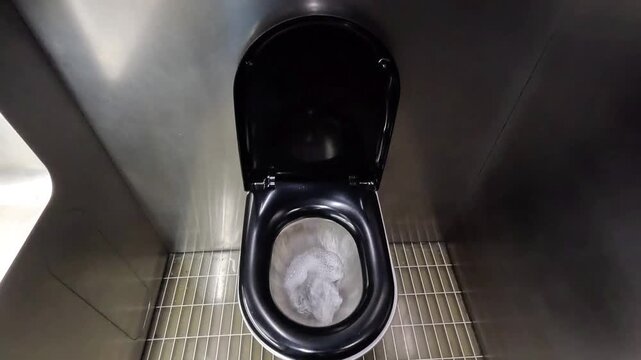 Modern black toilet bowl with water flushing in public restroom stainless steel stall.