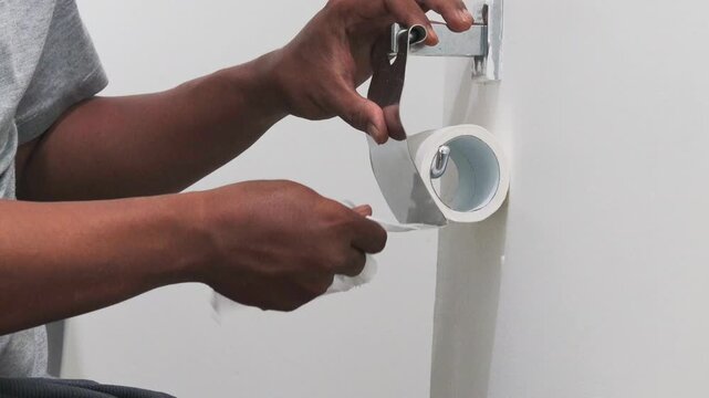 Close-up of Asian man hands unrolling toilet paper roll and tearing off a bit.