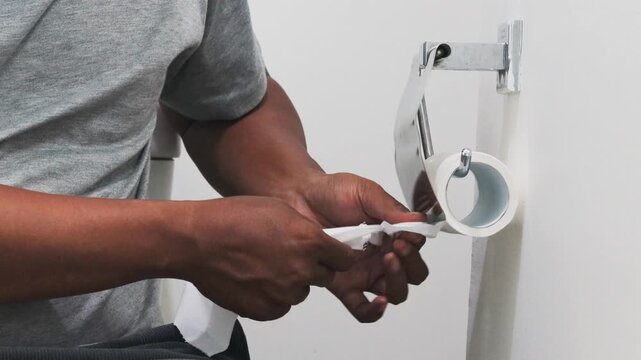 Close-up of Asian man hands unrolling toilet paper roll and tearing off a bit.