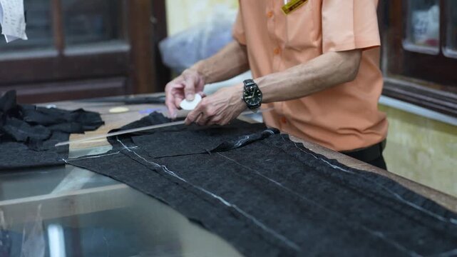 Traditional clothing manufactory in Vietnam, man employee at garment factory workshop making marks with crayon on textile pants patterns before sewing, working process of national clothing production.