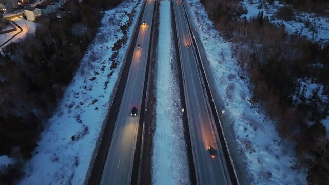 Drone aerial view of continuous traffic flow on a snowy highway during blue hour after sunset. Trucks and cars move steadily with bright headlights cutting through winter darkness, Nova Scotia, Canada