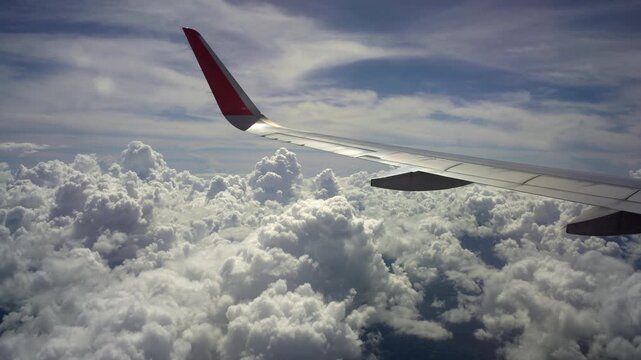 View from airplane window showing wing above dramatic clouds in the sky during flight travel scene with bright daylight and soft cloudscape