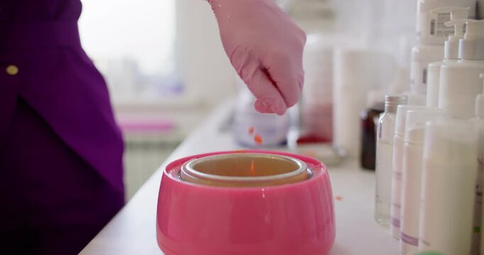Wax grains. Skin care. Unrecognizable female hand in pink latex glove using orange product granules for melting in heater preparation for professional cosmetic procedure.