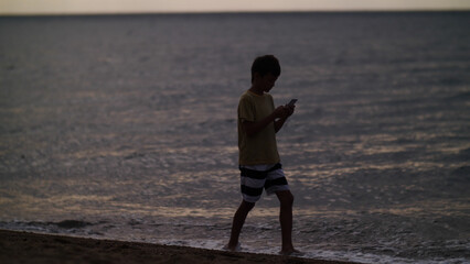 traveler boy walk on the beach and using smart phone in the evening, chats, write sms. Using a portable handheld device on summer vacation, internet. © vla