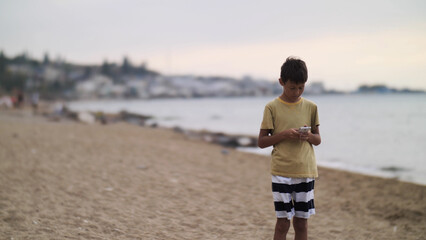 traveler boy walk on the beach and using smart phone in the evening, chats, write sms. Using a portable handheld device on summer vacation, internet. © vla