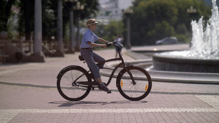Fototapeta premium happy boy in a hat rides a bike in the city park against the background of a fountain, have fun and enjoy