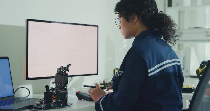 Young African American female robotics engineer coding AI software for robotic arm in laboratory. Embedded system, firmware testing, machine learning, industrial automation, future technology concept.