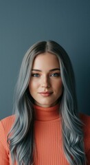 Fototapeta premium Serene young woman with long silver blue hair posing in coral turtleneck sweater for beauty editorial and fashion collection against muted blue studio background