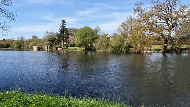 Peaceful countryside house by river with reflections in spring. Scenic rural floodgate house beside calm river with trees and reflections on a sunny spring day