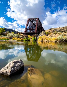 Rusty metal bridge over a reflective river with rocks in foreground and blue sky