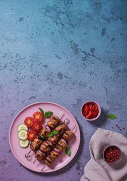 Grilled meat lula kebab skewers served on a pink plate with fresh vegetables over a blue textured background for a culinary blog or restaurant menu with copy space