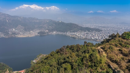 Fototapeta premium The beautiful blue lake is surrounded by mountains. Green forest on the slopes. A city in a valley. Snow-capped peaks of the mountains, azure sky. Pokhara. Lake Phewa. The Himalayas. Annapurna.