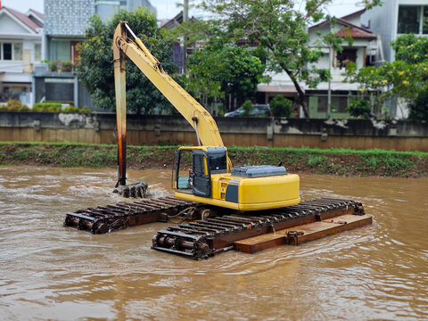 Amphibious excavator in yellow, mounted on pontoons with track attachments, dredging river sediment for waterway maintenance and flood control operations in shallow aquatic environments
