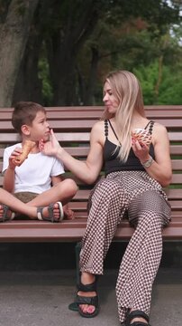 Vertical video. Bench leisure. Family eating outside. Happy laughing mother smiling young son together enjoying snack in green park.