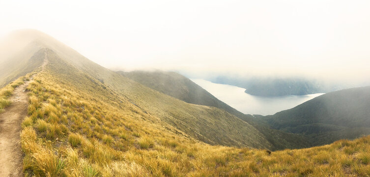 Misty mountain ridge hiking trail overlooking Lake Manapouri - Scenic panoramic landscape of New Zealand Southern Alps with golden grass, fog, and remote wilderness adventure