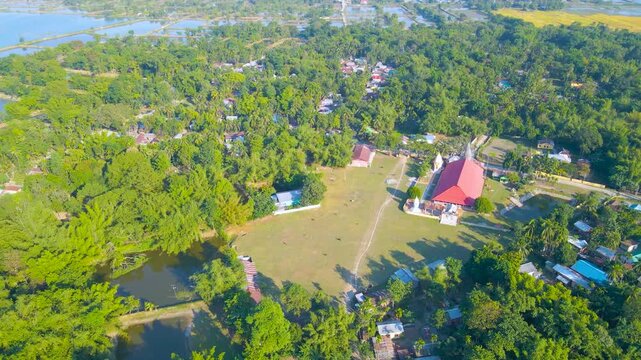 Drone shot of red-roof building with surrounding houses, fields, trees, and water showcasing rural settlement in Byaskuchi, Assam.