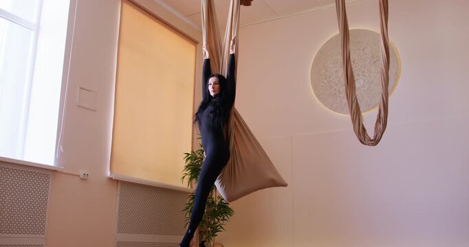 Fly yoga. Female stretching. Athlete doing acrobatic exercise in split pose supported by fabric hammock wrapped around leg in gym.