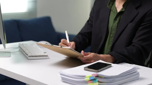 Professional businessman working on computer project with paperwork, analyzing data on a computer and taking notes on a clipboard with a stack of documents nearby.