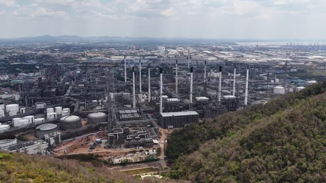 Wide aerial perspective of a massive oil refinery complex, featuring storage tanks, pipelines, and smokestacks adjacent to a forested hill with a sprawling industrial port and city in the distance