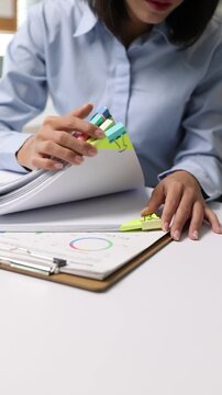 Asian female employee sorting through a stack of paperwork, adding colorful sticky tabs to organize files at her office desk.