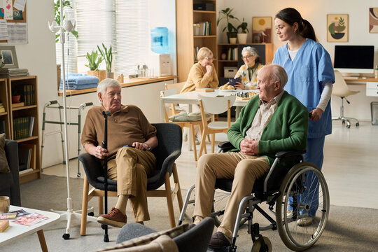 Wide shot of senior men interacting with female caregiver in nursing home, showing assisted living support and mobility care. Useful for eldercare marketing, healthcare services, caregiving campaigns