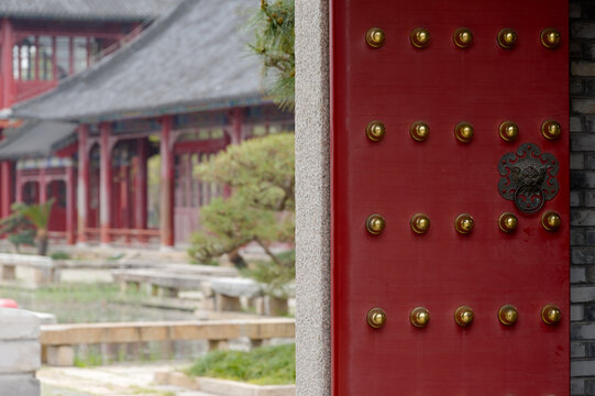Close-up of a traditional Chinese red temple door with decorative brass studs and an ornate knocker, partially revealing a blurred historic courtyard and wooden temple architecture, evoking cultural