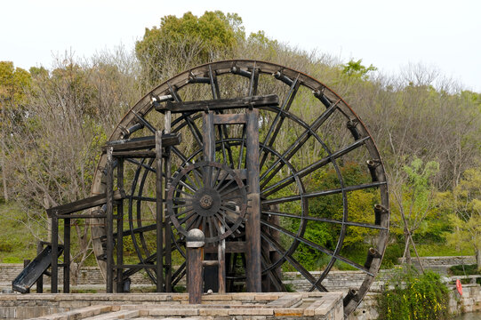 Large historic wooden waterwheel standing beside a stone terrace, framed by trees and greenery in a peaceful park landscape, rustic architecture and traditional engineering evoke heritage and calm.