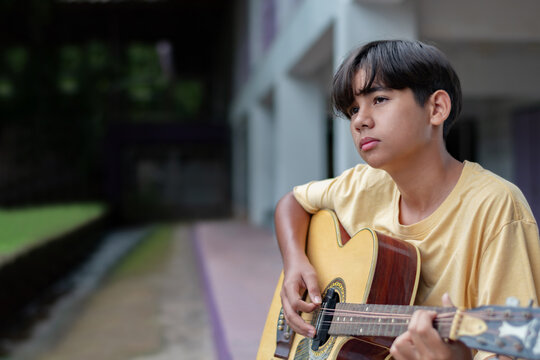 Teen boy playing acoustic guitar outdoors, wearing a yellow t-shirt, looking thoughtful. Creative youth expression and musical lifestyle in a casual setting.