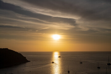 Obraz premium Golden sunset over the Andaman Sea from Windmill Viewpoint in Phuket, with sailboats drifting on calm water and warm reflections stretching across the horizon.