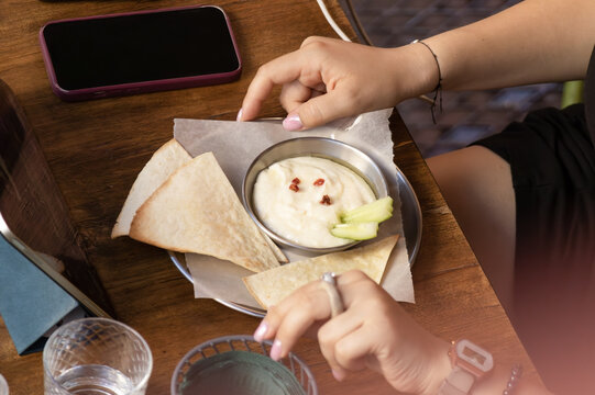 Small plate with flatbread, creamy dip, and cucumber slices sits on wooden table alongside phone, water glass, and hands in relaxed dining setting. Scene conveys informal atmosphere, texture, and ever