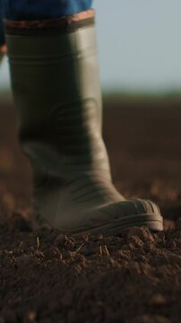 Close-up of sturdy green rubber boots walking on dry cultivated soil in an open agricultural field, symbolizing farming, outdoor labor, and rural countryside work