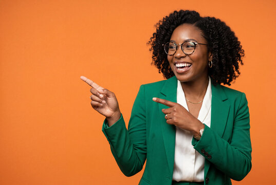 Smiling business woman in a green blazer pointing aside against a bright orange studio wall