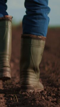 Close-up of a person in green rubber boots walking along a freshly plowed soil path on a farm field, highlighting rural work, agriculture, and outdoor lifestyle