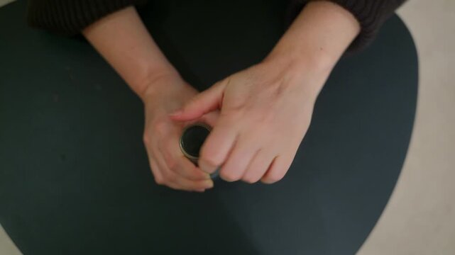 Top view of a person's anxious hands wringing and fidgeting on a dark table. The individual then holds a small round container, a possible coping mechanism for stress, worry, or nervousness