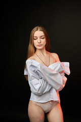 A young woman poses in a white off-the-shoulder shirt on a black background. The soft lighting highlights her relaxed and confident expression. She is wearing minimal underwear, which adds to her sens © SergeiPh