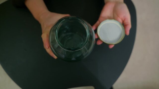 Top down view of female hands opening and closing green glass jar with golden lid. Person unscrews and screws cap on empty container on dark green surface. Concept of storage and preservation