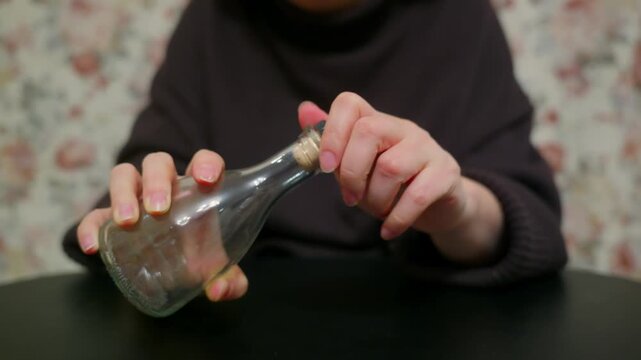 Close up shot of woman's hands putting cork into empty glass bottle. Person seals small transparent flask with wooden stopper at dark table. Simple, focused action