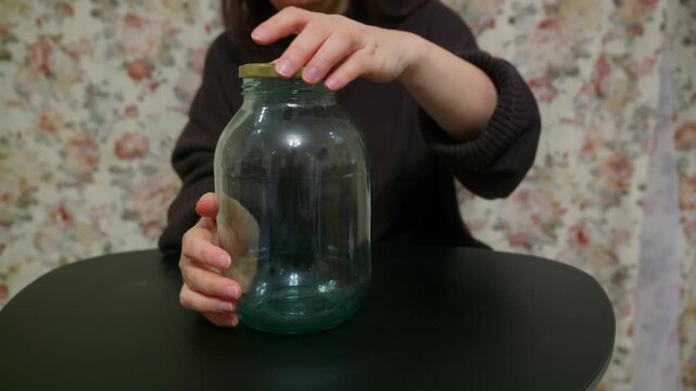 Close up of female hands unscrewing and opening large empty glass jar. Person twists golden lid on canning jar standing on black table. Domestic scene with floral background. Concept of preserving