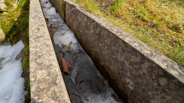 Close view of a narrow concrete drainage channel with melting snow, thin ice, and trapped leaves, capturing seasonal runoff and wet surface texture in early spring.