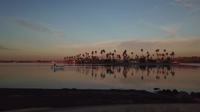 Static drone shot at Mission Bay at 6:30AM during sunrise with a boat in the water and birds flying across a peaceful morning sky.