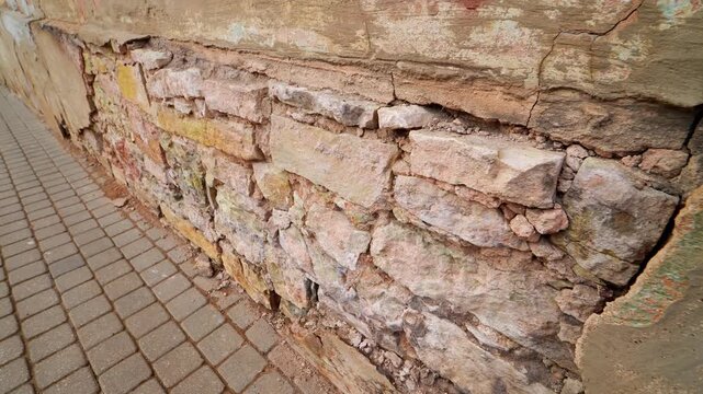 Close-up of deteriorating stone foundation wall with cracked plaster and exposed masonry beside sidewalk, showing weather damage and poor repair on old building facade.