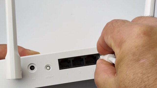 A hand is inserting a white ethernet cable into a port on a white router. The router has multiple ports labeled 1, 2, 3, and 4.
