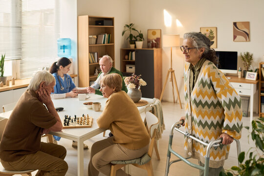 Senior Caucasian woman with disability using walker while elderly group socializing and playing chess in care center. Ideal for senior care, assisted living, rehabilitation marketing